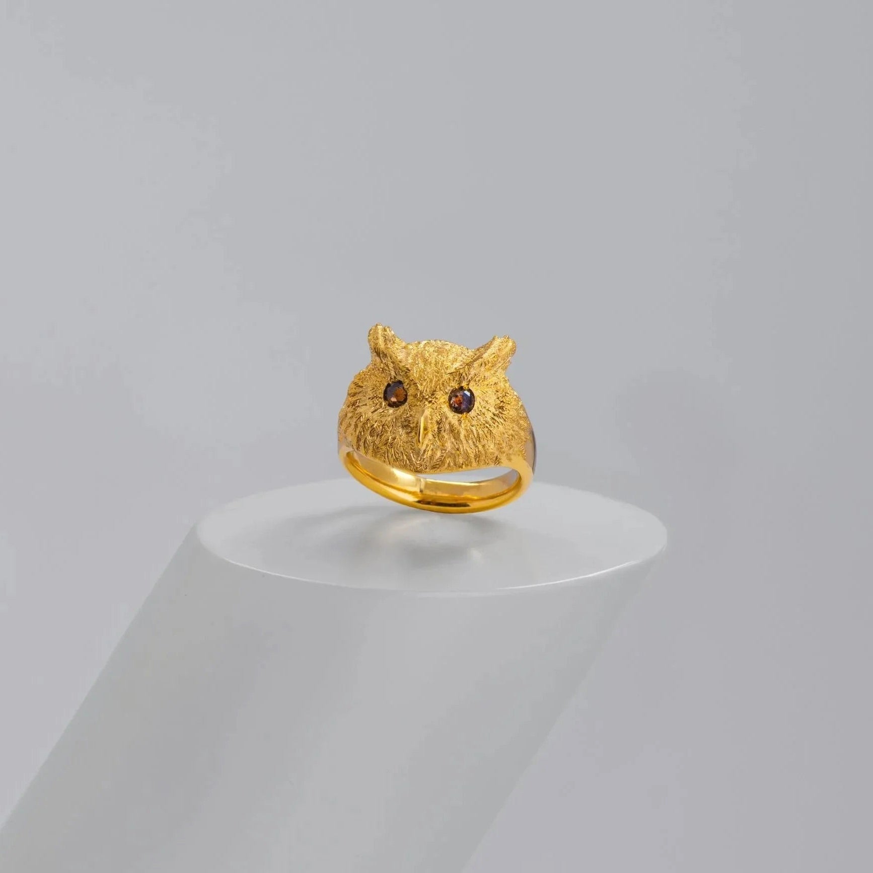 A gold owl ring with textured feather-like detailing and dark eyes, displayed on a white cylindrical pedestal against a light gray background.