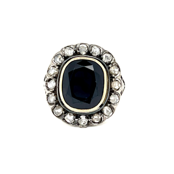 A vintage-style oval ring featuring a large dark blue faceted center stone surrounded by a halo of small clear stones in a silver-toned setting with a gold inner border, shown on a plain white background.