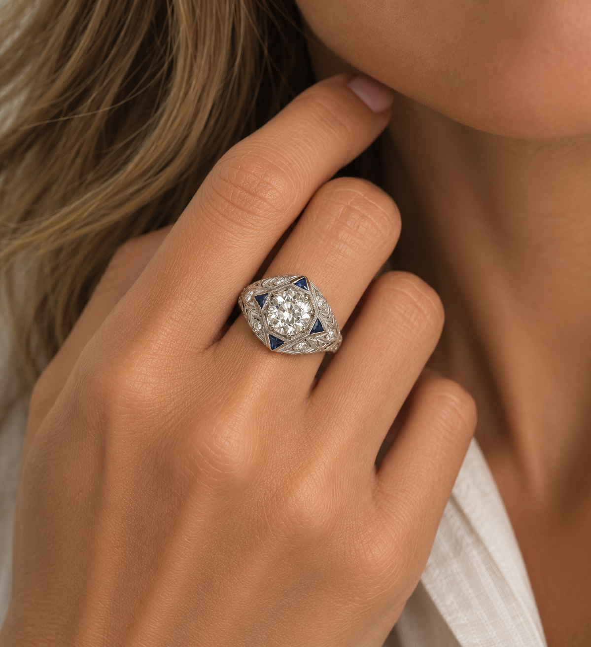 A close-up lifestyle shot of a woman's hand wearing a vintage-style diamond ring with blue accents on a white background.