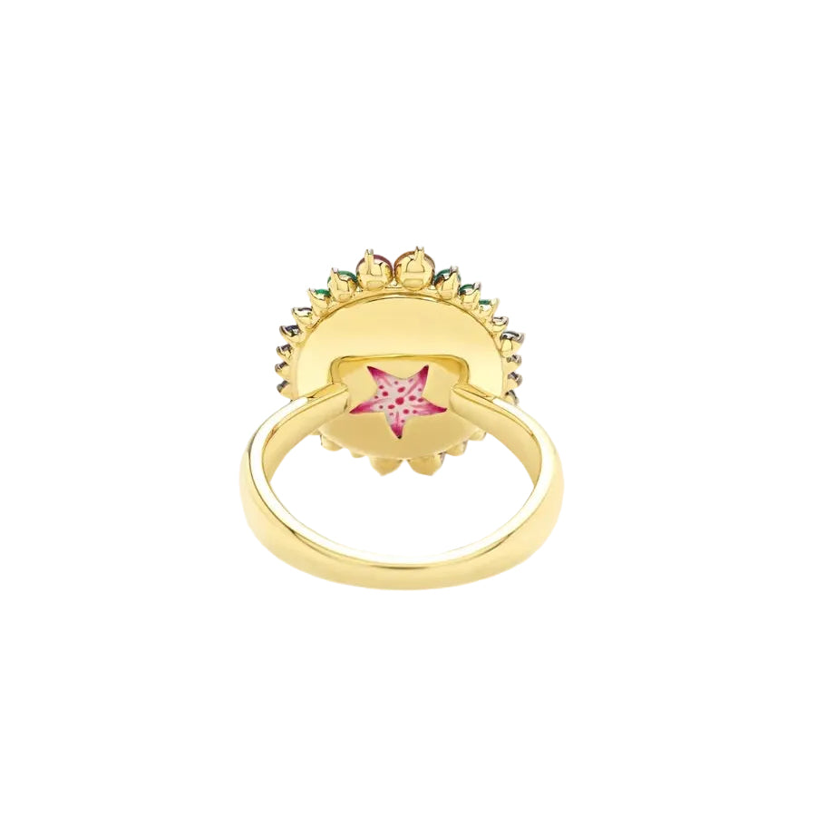 A gold ring featuring a decorative sunburst design with small green and yellow stones, and a central pink star-shaped gemstone, shown on a light beige background.