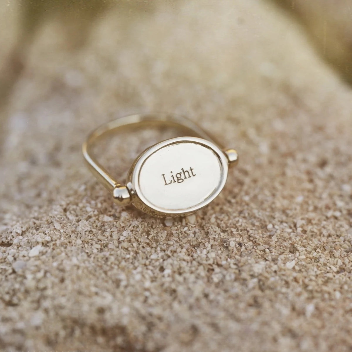 A gold signet ring with an oval face engraved with the word 'Light', resting on a sandy surface.