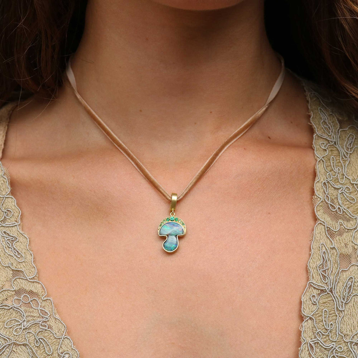 A close-up of a person's neck and chest wearing a delicate gold necklace with a mushroom-shaped pendant featuring a light blue iridescent center and green accents, paired with a beige lace top.