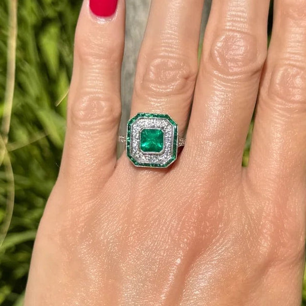 Hand with red nail polish wearing a green emerald ring against a blurred green background
