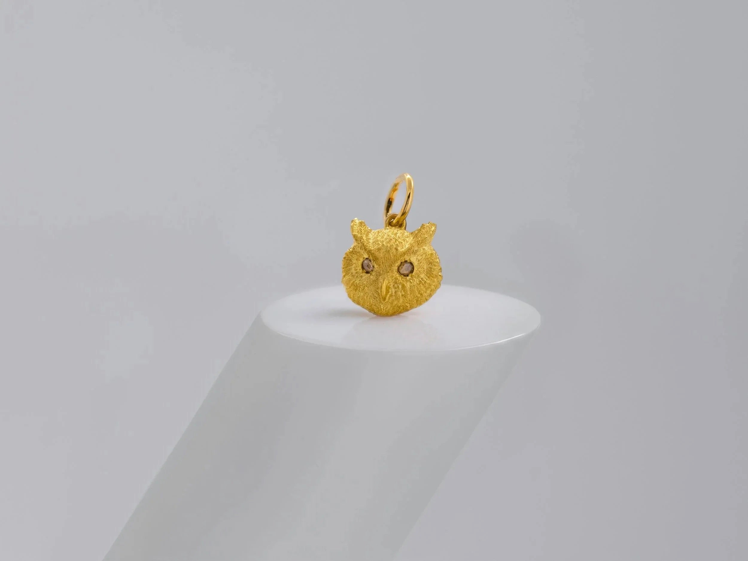 A textured gold pendant shaped like a small owl head with tiny dark eyes, displayed on a white cylindrical pedestal against a soft light gray background.