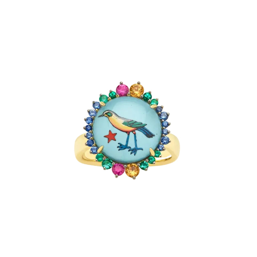 A gold ring featuring a round light blue cabochon with an illustrated bird and red star, surrounded by a halo of blue, green, and pink gemstones on a light beige background.