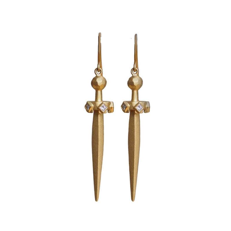 A pair of gold dagger-shaped drop earrings with small clear gemstone accents on the crosshairs, hanging from a dark horizontal bar against a soft gray background.