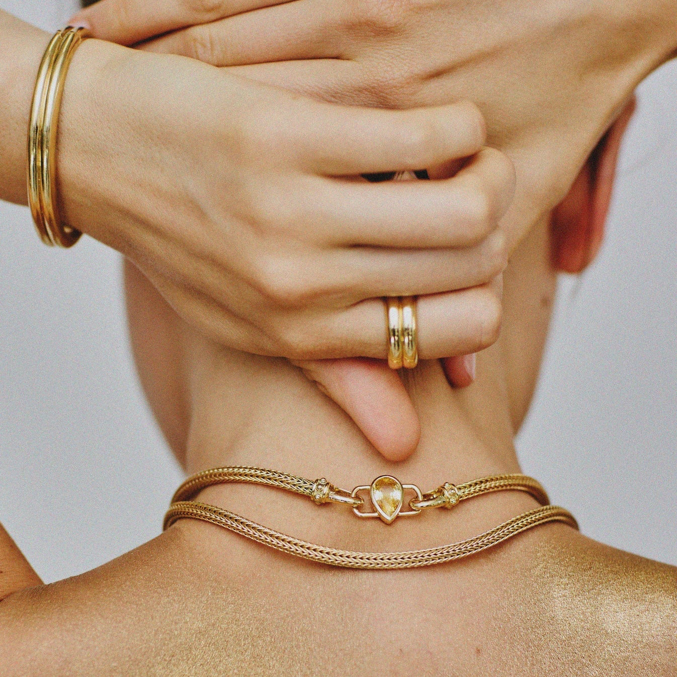 Close-up of a person wearing gold jewelry including a bracelet, ring, and necklace on a neutral background.