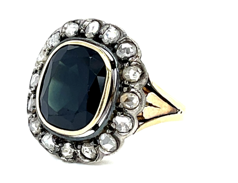 An antique-style ring featuring a large oval-cut dark blue stone in a gold bezel, surrounded by a halo of small clear stones on a white background.