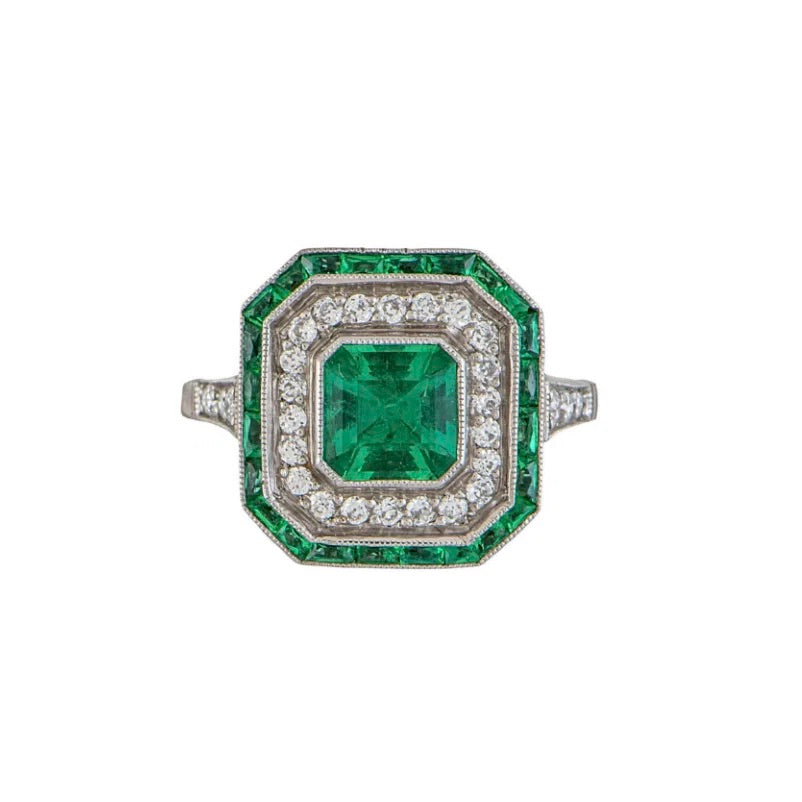 An antique-style emerald-cut emerald ring with a silver-toned metal setting, featuring a central emerald green stone surrounded by a double halo of small round diamonds and a border of emerald-cut green stones, set against a plain white background.