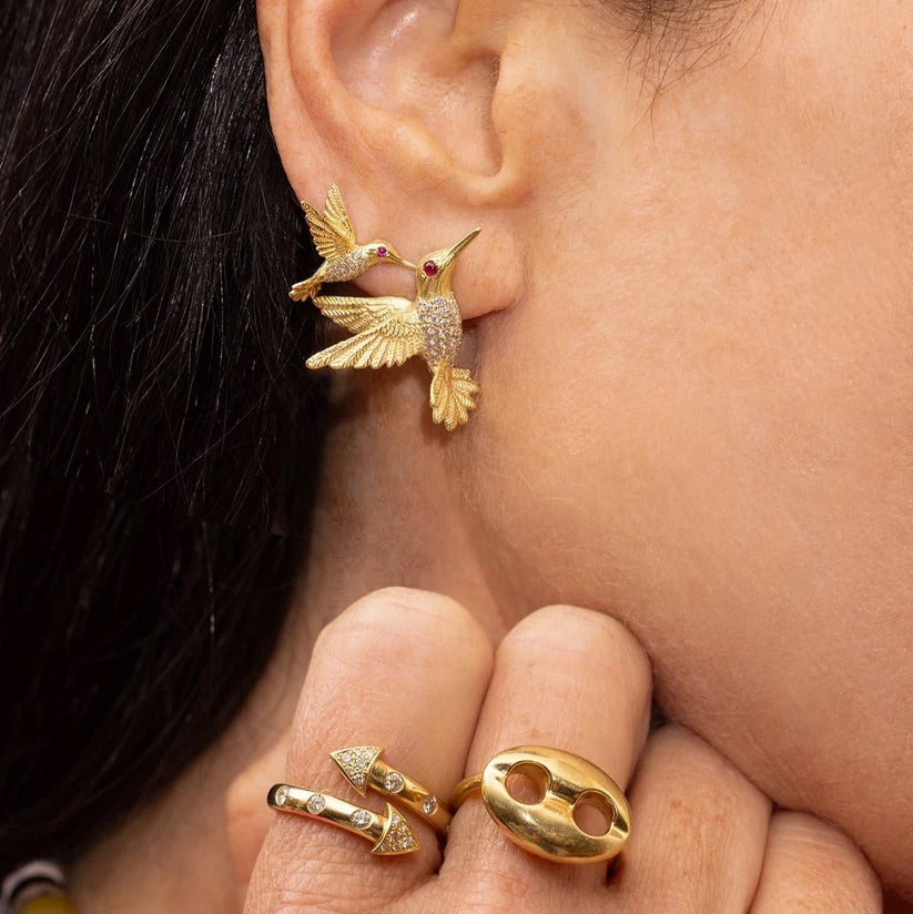 A close-up of a person's ear wearing a gold bird earring, with two small gold birds perched on the ear. The person is also wearing two gold rings on their fingers.