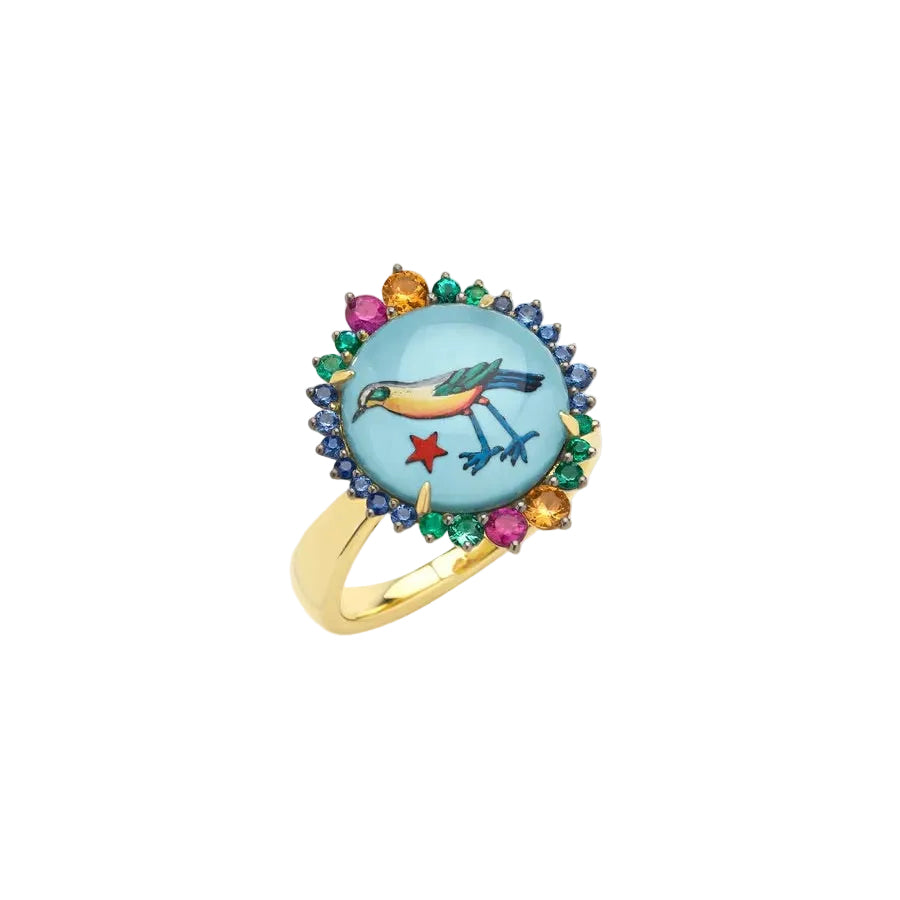 A gold ring featuring a round light blue enamel inlay with a painted bird and small red and yellow accents, surrounded by a border of blue, green, and pink gemstones on a light beige background.