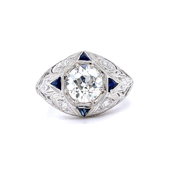 An antique-style diamond ring with a large round center stone, surrounded by small diamonds and accented with dark blue triangular gemstones on a silver-toned filigree band, shown on a white background.
