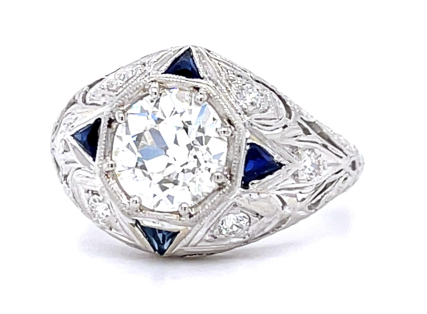 An ornate Art Deco style diamond ring featuring a large round brilliant-cut center stone, surrounded by small diamonds and accented with dark blue triangular gemstones on a silver-toned metal setting, shown on a white background.