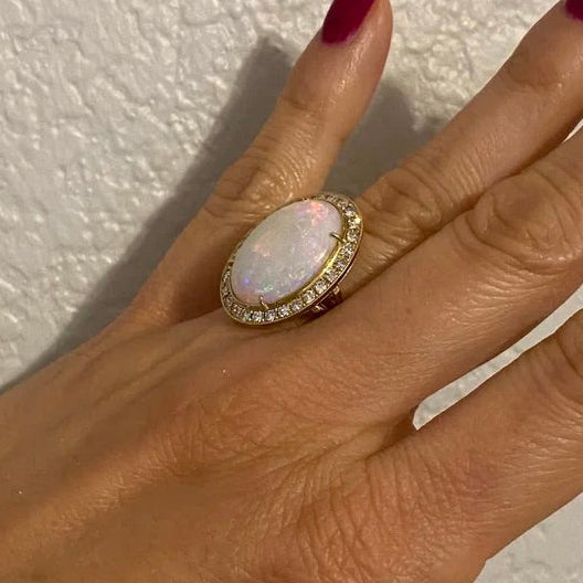 A close-up of a hand with dark burgundy nail polish wearing a gold ring with an oval white opal and small clear stones, set against a textured off-white wall.