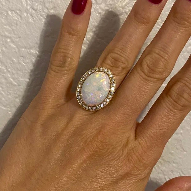 A hand with dark red nail polish wearing a large oval opal ring with a halo of small clear stones, set against a textured off-white wall.