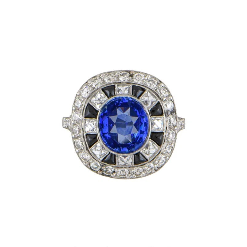 An Art Deco style ring featuring a round brilliant-cut blue sapphire center stone, surrounded by a halo of small clear diamonds and black triangular accents, set in a silver-toned metal band.