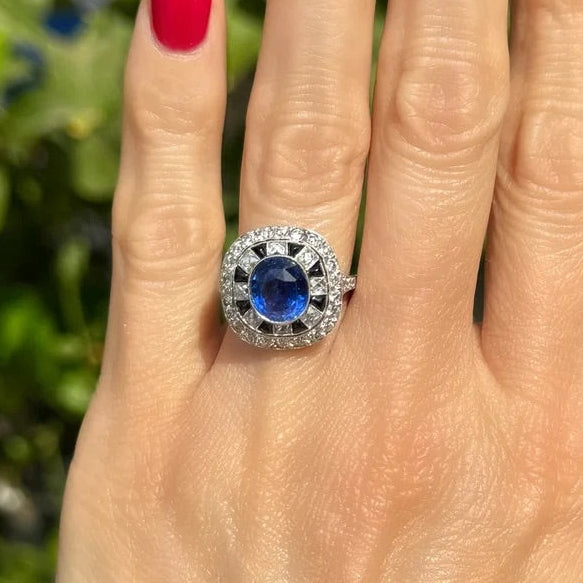 A close-up of a hand with bright pink nail polish wearing a silver ring with a round blue gemstone surrounded by small clear stones, set against a blurred green garden background.