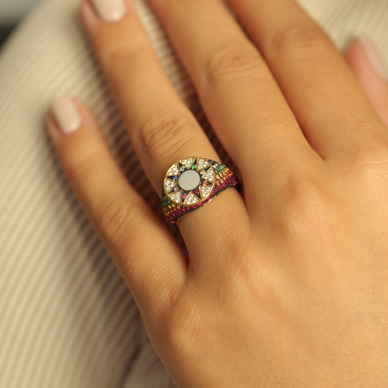 A close-up of a hand wearing a colorful ring with a round white center and multiple colored stones around it, resting on a ribbed cream fabric.