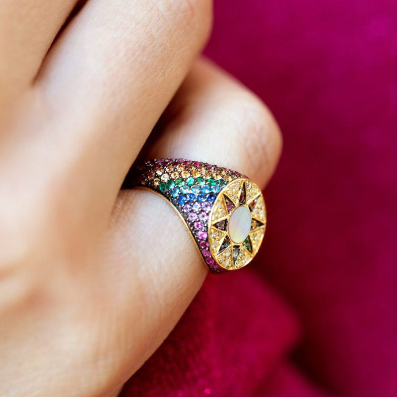 A close-up of a hand wearing a gold signet ring adorned with multicolored pavé gemstones and a central iridescent stone, set against a vibrant magenta fabric background.