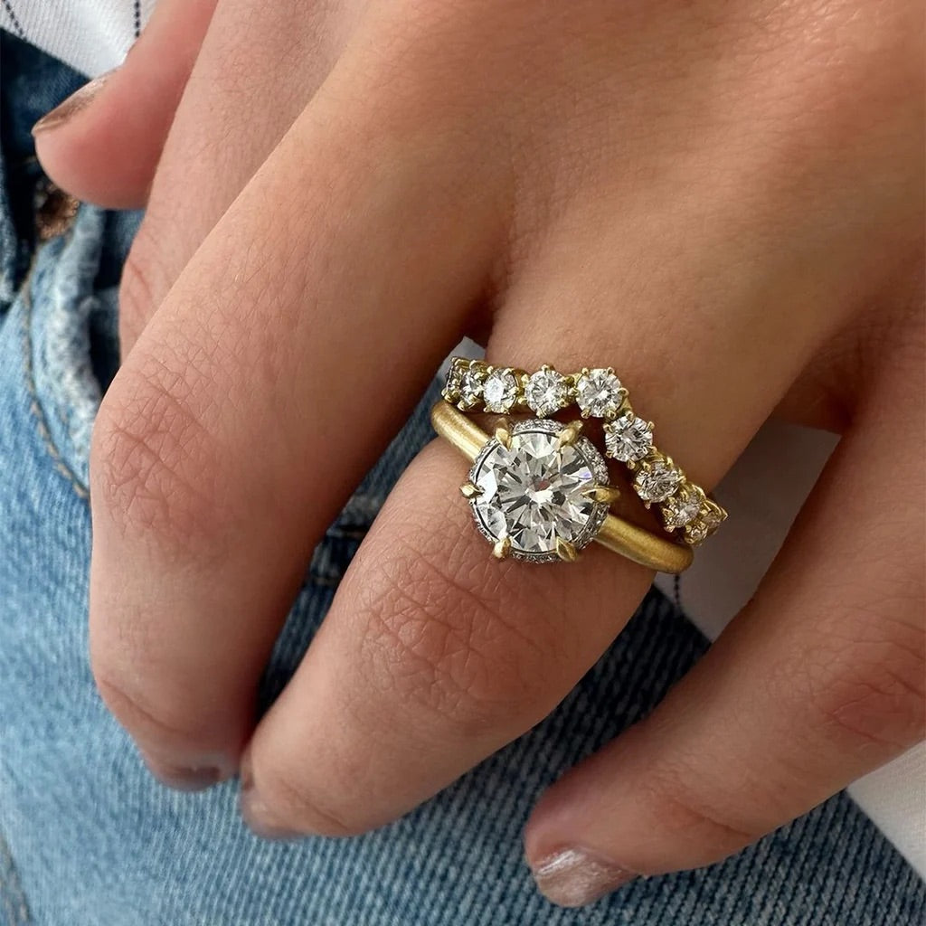 A close-up of a hand wearing a yellow gold engagement ring with a large round clear center stone and a matching yellow gold band with small clear stones, set against blue denim.