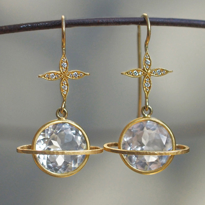 A pair of gold dangle earrings with a four-pointed star motif set with small clear stones, hanging from a dark metal rod. Each star is connected to a large, round, faceted clear gemstone in a gold bezel setting with a thin gold ring around it, set against a soft, out-of-focus gray background.