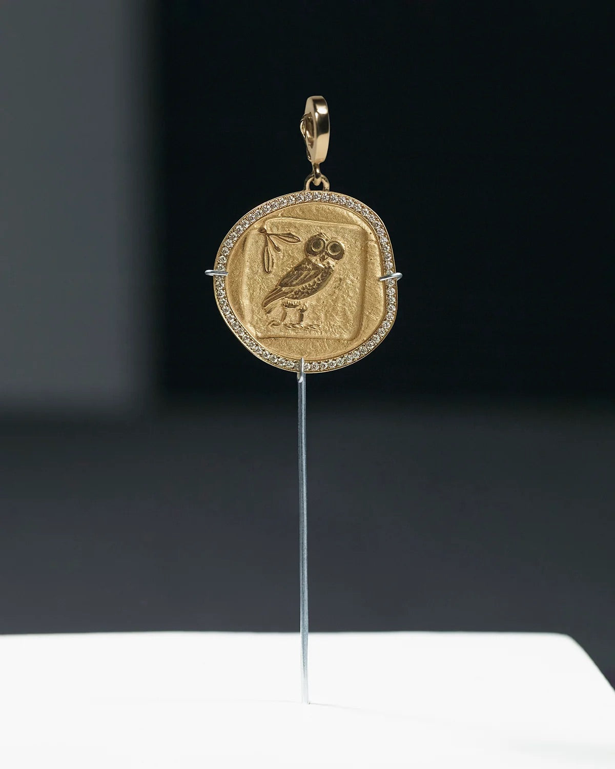 A gold pendant shaped like an ancient coin, featuring an owl motif in the center, displayed on a thin stand against a dark background.