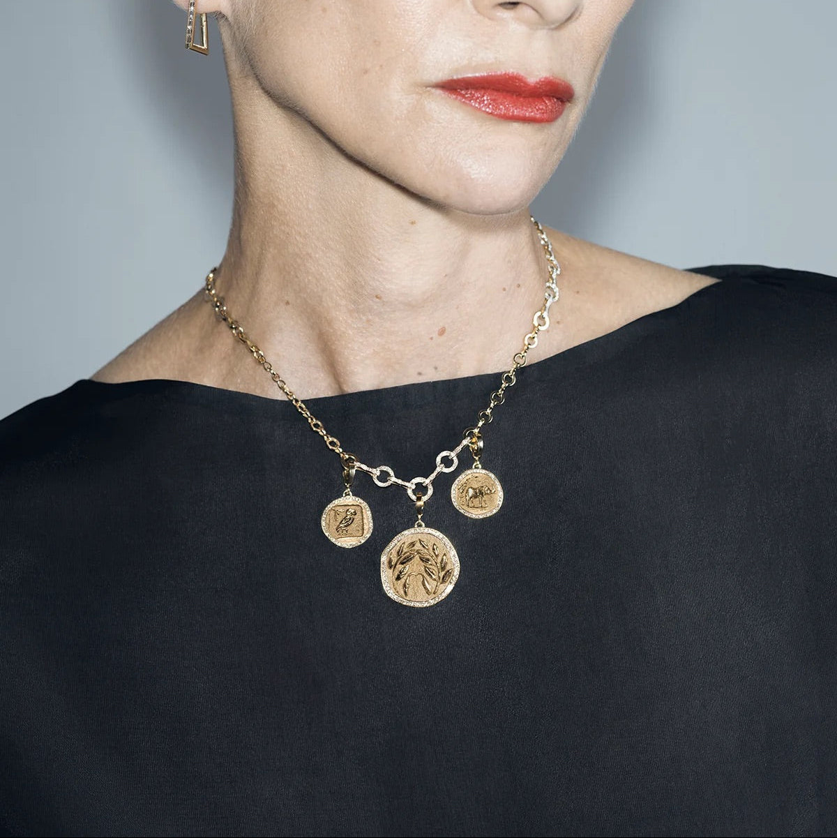 A close-up portrait of a woman wearing a gold coin-link necklace with three dangling circular medallion charms, a small matching hoop earring, and a black top against a light gray background.