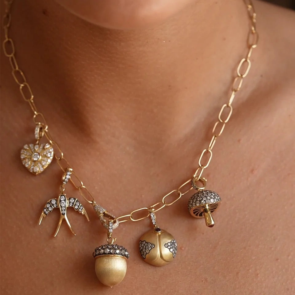 A close-up of a gold chain necklace worn by a model, featuring five dangling charms: a heart-shaped flower with clear stones, two silver-and-gold birds, a mushroom with clear stones, and an acorn with a cream-colored pearl-like center and clear stones.