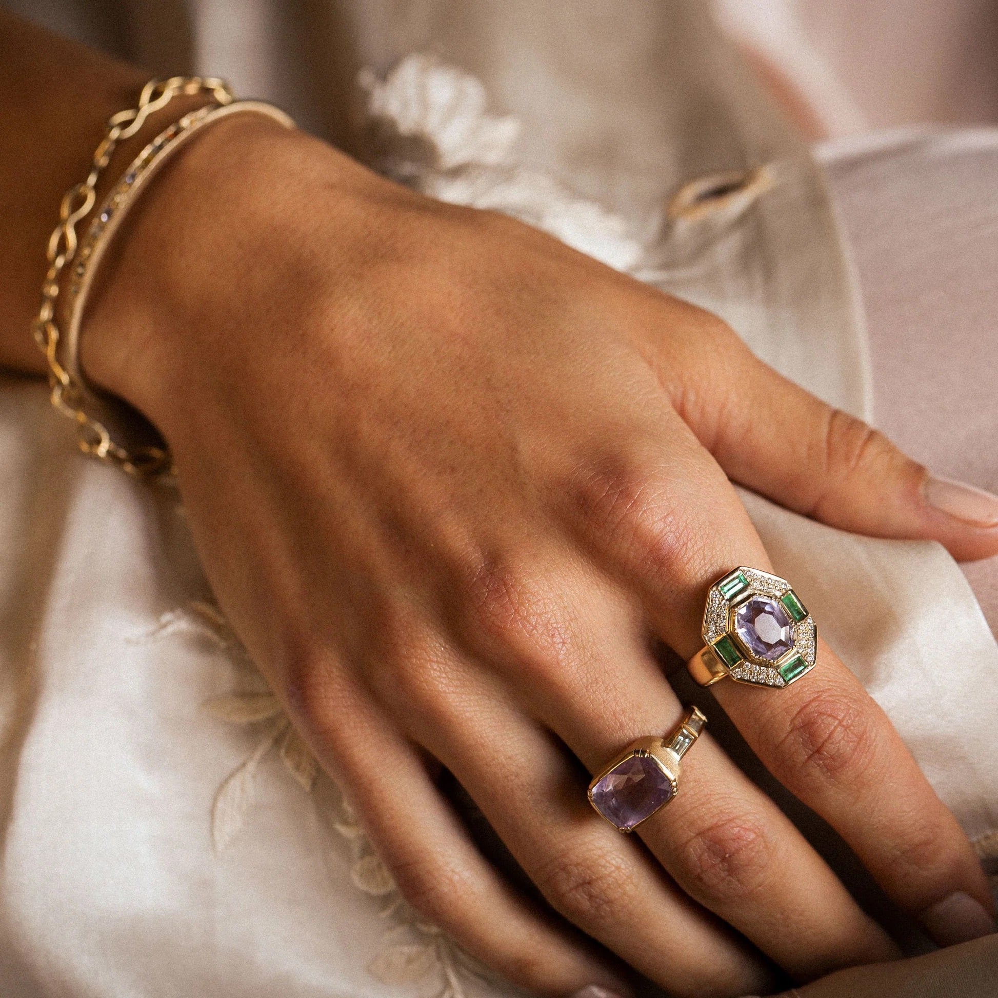 A close-up of a hand wearing two gold rings with purple gemstones and green accents, along with a gold chain bracelet, resting on a light cream fabric.