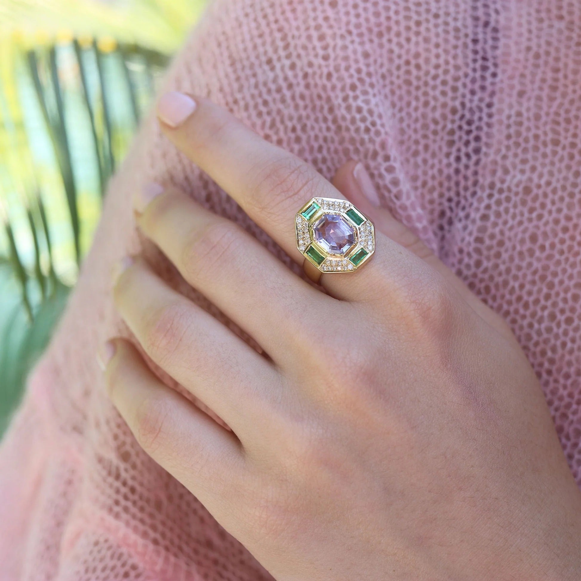 A gold ring with a central lavender-purple oval gemstone and green accent stones, worn on a person's finger against a pink textured fabric background.