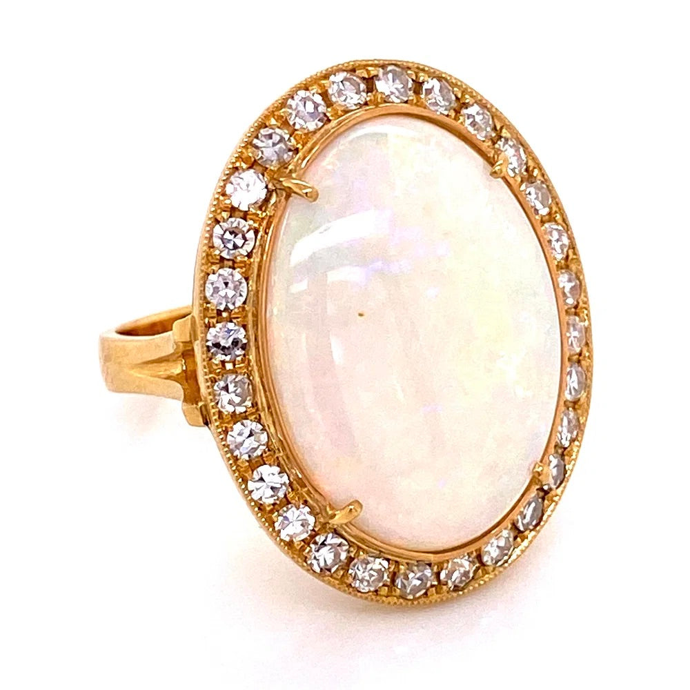 A yellow gold ring featuring a large oval white opal center stone surrounded by a halo of small clear round-cut diamonds, set against a plain white background.