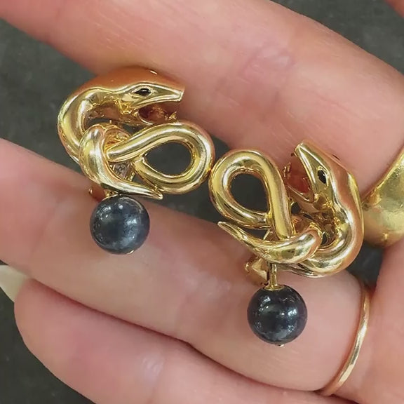 Gold snake-shaped earrings with black beads held in a hand.