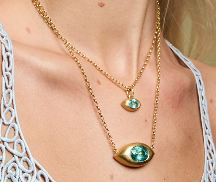 A close-up of a model wearing a double-layered gold necklace. The shorter chain features a small gold teardrop pendant with a green gemstone, while the longer chain features a larger gold eye-shaped pendant with a matching green gemstone.