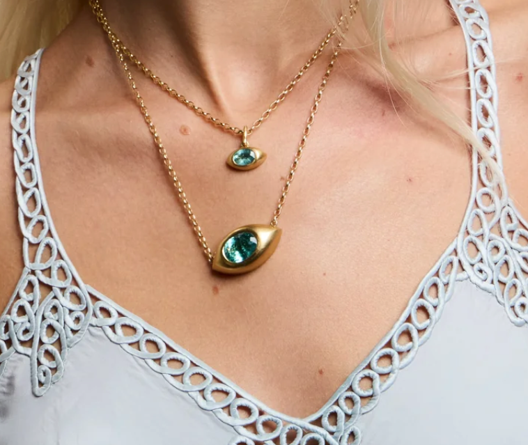 A close-up of a model wearing two layered gold necklaces with teardrop-shaped green gemstone pendants, styled with a white eyelet lace top.