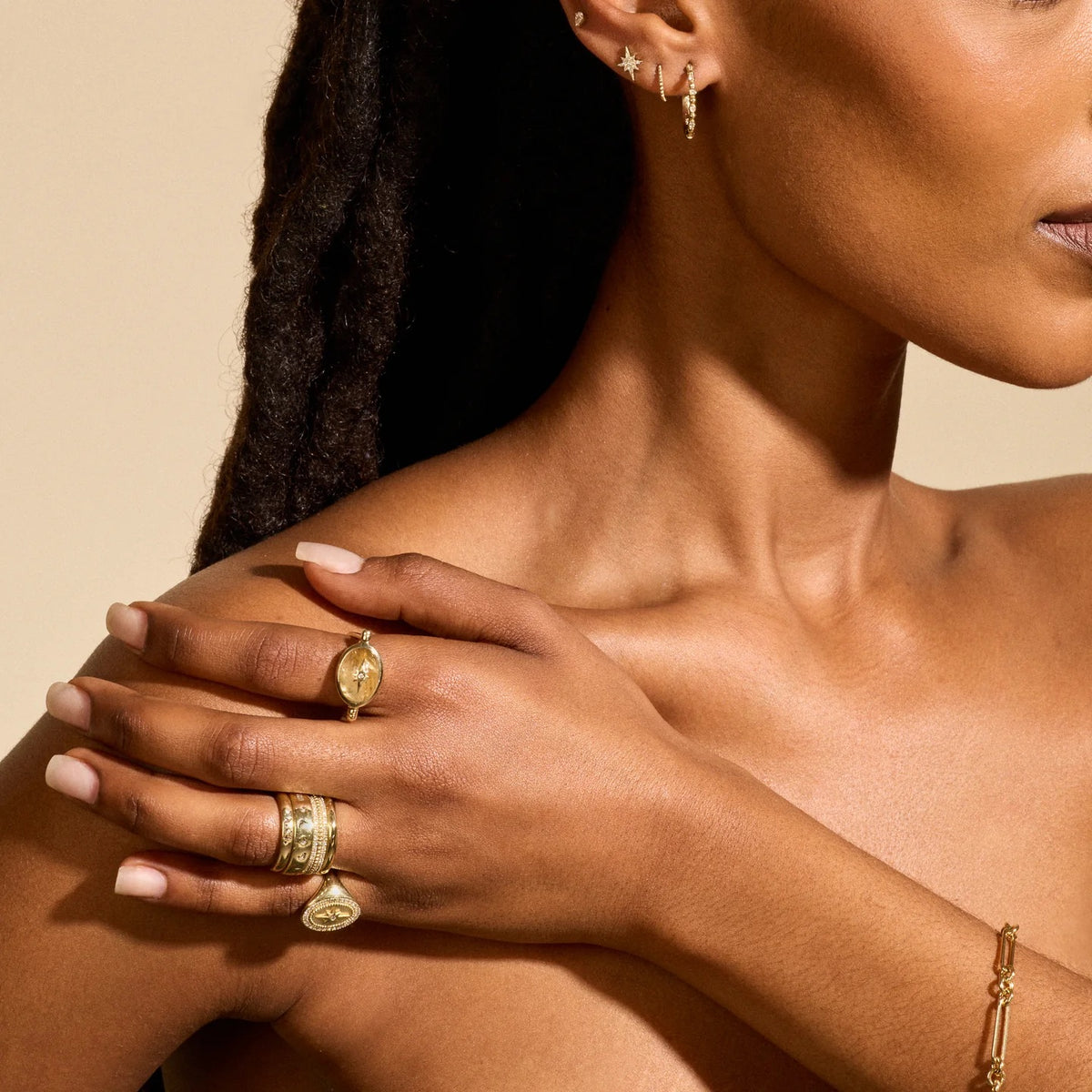A close-up lifestyle shot of a model wearing multiple gold rings on her fingers, including a textured band, a signet-style ring with a gold heart, and a small gold chain bracelet on her wrist, against a warm beige background.